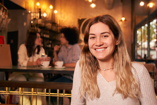 Copy space portrait of cheerful blonde girl at bar. Empowered European woman poses looking at smiling camera. Girlfriends having fun indoors. Three co-workers after shopping. Happy people.