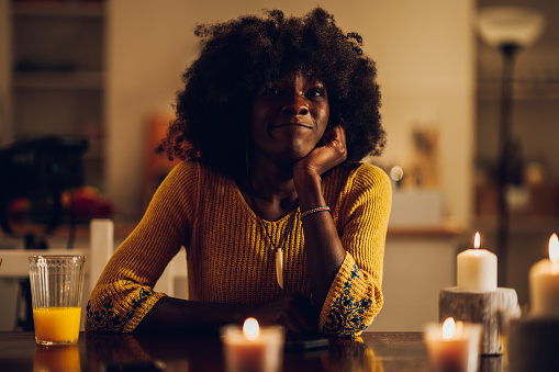 Portrait of an african american woman looking into the camera in the night while sitting at the table at home with candles on it. Black female with afro hair in a yellow knitted shirt. Copy space.