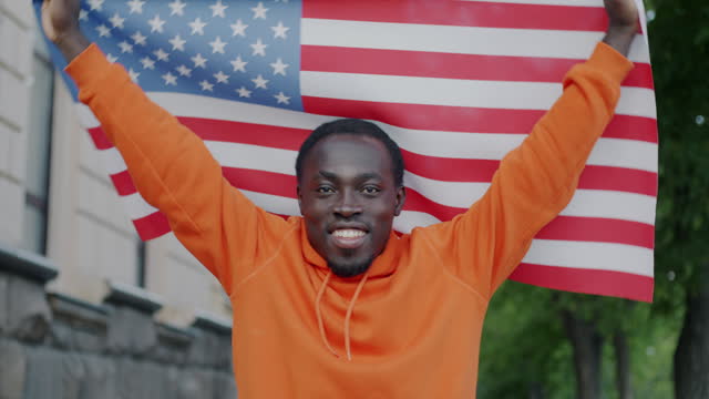 Slow motion portrait of joyful African American man waving US flag smiling looking at camera outdoors in city