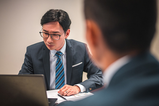 Waist up shot of a mid adult Asian male corporate employee using a laptop during a business meeting. He is wearing an elegant suit and tie.