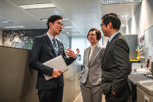 Three-quarter length shot of three finance executives standing and talking in a corporate office. They are happy and smiling. They are socializing at work.