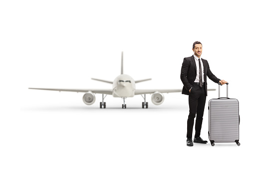 Full length portrait of a young man in a suit posing with a suitcase in front of an aircraft isolated on white background