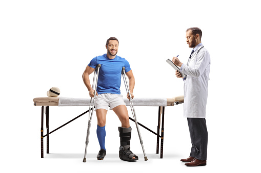 Football player with crutches sitting on a physical therapy bed and a doctor writing a document isolated on white background