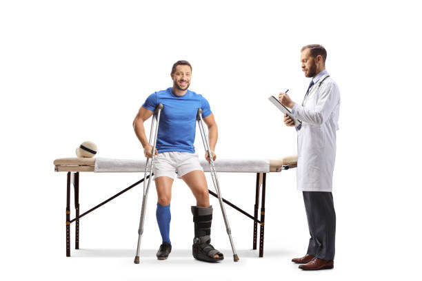 football player with crutches sitting on a physical therapy bed and a doctor writing a document - physical injury men orthopedic equipment isolated on white imagens e fotografias de stock
