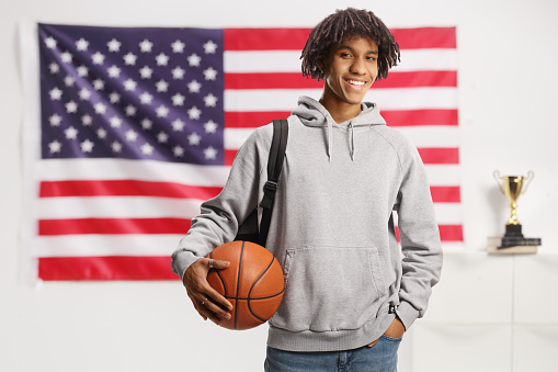 Young african american male with a backpack holding a basketball and smiling in front of a USA flag