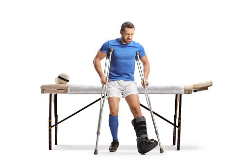 Male athlete with crutches sitting on a massage bed isolated on white background