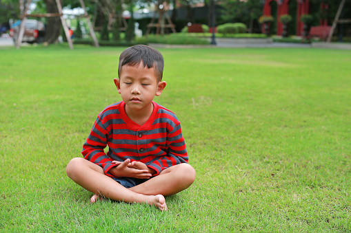 Asian little boy child practicing mindfulness meditation sitting on lawn in the garden.