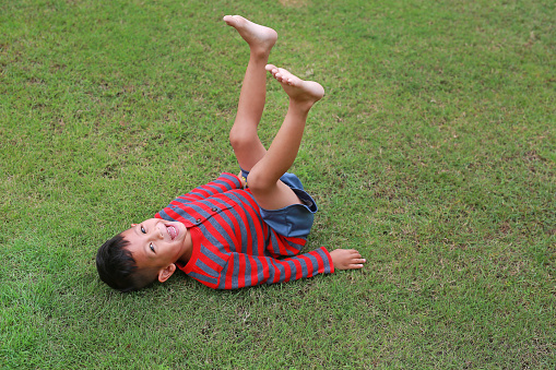 Smiling Asian little boy lying on green lawn with lift up his legs. Kid lies on grass with looking camera.