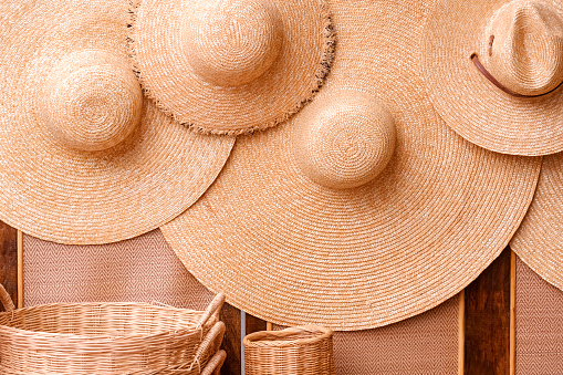 Group of straw floppy hats hanging on woven partition with wicker baskets showing for sale in street market at Thailand, Wicker products background.