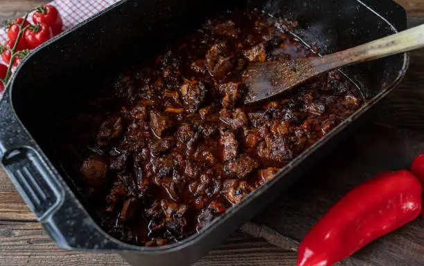 Delicious oven braised pork cut into cubes. Cooked with tomatoes, bell peppers, onions, garlic and herbs. Served hot and ready to eat in a roating pot on rustic and wooden table background from above.