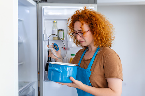 Red-haired woman meticulously inspects the fridge for spoiled food. Her discerning eyes and cautious demeanor ensure freshness, capturing the essence of responsible and mindful food management