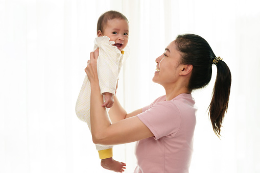 cheerful mother holding and lifting with her infant baby on a window background