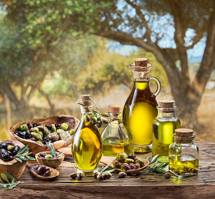 Bottle of craft farm olive oil stands on a wooden table, behind is an olive garden in a slightly bokeh, sunny day.