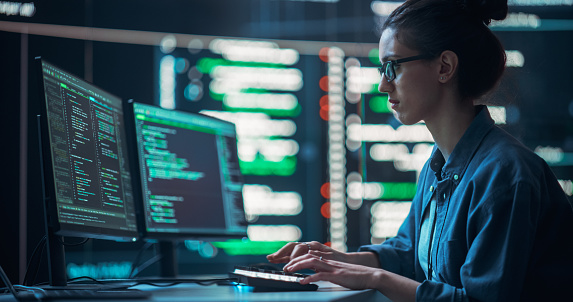 Medium Shot of a Woman Working as a Developer, Surrounded by Big Screens Displaying Lines of Code in a Monitoring Room. Female Programmer Using Desktop Computer, Analysing Data, Creating AI Software