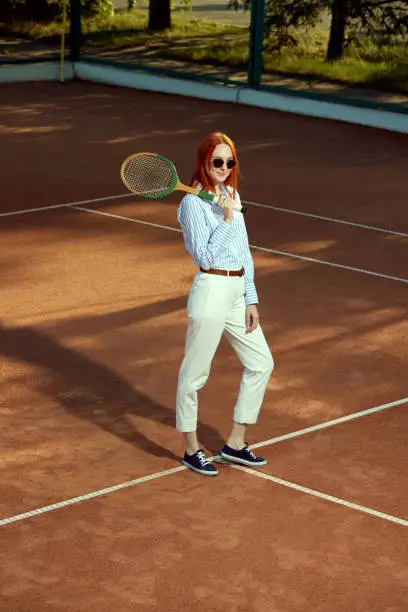 Full-length top view image of redhead young woman in stylish casual clothes and sunglasses posing on opendoor tennis court. Concept of sport, active lifestyle, hobby, casual fashion, leisure time, ad