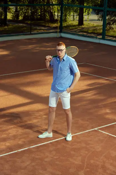 Full-length top view image of handsome redhead young man in stylish casual clothes and glasses posing on opendoor tennis court. Concept of sport, active lifestyle, hobby, fashion, leisure time, ad