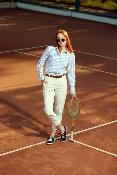 Full-length top view image of beautiful redhead young woman in stylish casual clothes and sunglasses posing on opendoor tennis court. Sport, active lifestyle, hobby, fashion, leisure time, ad concept