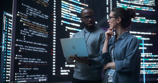 istock Portrait of Two Diverse Developers Using Laptop Computer, Discussing Lines of Code that Appear on Big Screens Surrounding Them. Male and Female Programmers Creating a Software Together and Fixing Bugs 1518860626