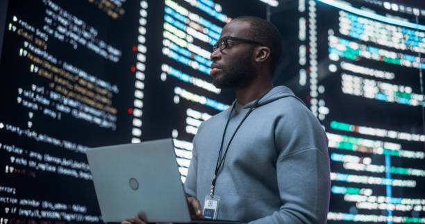 istock Portrait of Young Black Man Working on Laptop Computer, Looking at Big Digital Screen Displaying Back-end Code Lines. Professional Programmer Developing a Big Data Interface Software Project Concept. 1518861143