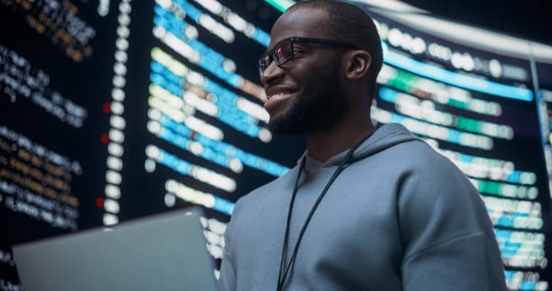 istock Portrait of Young Black Man Working on Laptop Computer, Looking at Big Digital Screen Displaying Back-end Code And Smiling. Professional Programmer Developing a Big Data Interface Software Project. 1518861153