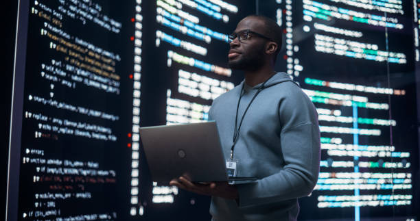 istock Portrait of Young Black Man Working on Laptop Computer, Looking at Big Digital Screen Displaying Back-end Code Lines. Professional Programmer Developing Big Data Interface Software Project. 1518862695