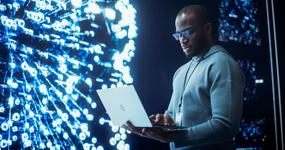 Portrait of Young Black Man Working on Laptop Computer, Looking at Big Digital Screen Displaying Neural Network Visualisation in 3D. Professional Data Specialist Analysing User Information