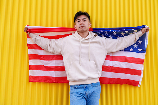 Vibrant photo of a Chinese man proudly posing with a US flag, set against a yellow wall, creating a striking and colorful composition.