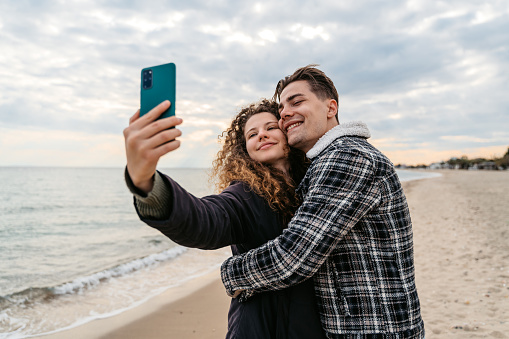 Beautiful young couple using a smart phone together on the beach in Nea Flogita, Greece. Taking selfies and filming.