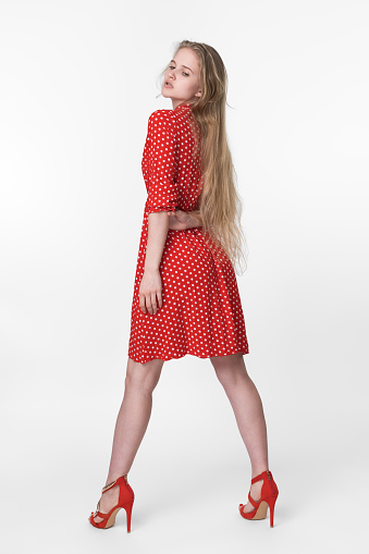 Stylish blonde woman with long hair dressed in summer red polka dot dress, red shoes standing in full length and poses. European female 21 years old looking down. Studio shot on white background. Part of series