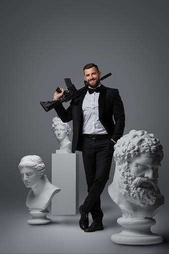 Fashionable man with a well-groomed beard wearing a black suit and bow tie, holding a gun standing confidently and smiling, posing with three antique statues on a gray background