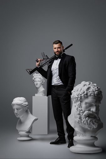 Stylish man with a well-groomed beard dressed in a luxurious black suit and bow tie, holding an automatic rifle striking a confident pose among three antique statues against a grey background