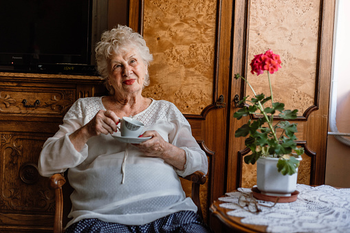 The woman is sitting on the chair in the living room, holding a cup of coffee and thinking. The senior woman drinking coffee/tea at home.