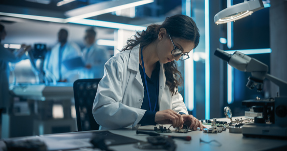 Proffesional Female Engineer Working on Electronic Circuit Boards. On Blurred Background Industrial Robotics Specialists in Lab Coats Working on a Mobile AI Robot in a Tech Factory Facility.