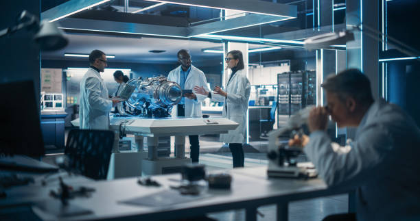 istock Young Team of Multicultural Engineers Use Blueprints, Tablet, Laptop Computers and Microscope to Analyze and Discuss How Electric Turbine Motor Works. High-Tech Research Laboratory with Equipment 1518983038