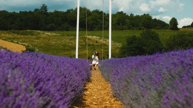 Girl sitting on a white wooden swing or bench in a lavender field of flowers. A woman walks and enjoys the floral glade and summer nature. Closeup. Natural cosmetics concept. France, Provence.