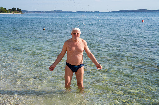 Senior men a Parkinson Disease patients in sea water Playing with water. He is trying to cool down before entering the water. It is early spring near Peroi, Istrian coast in Croatia.