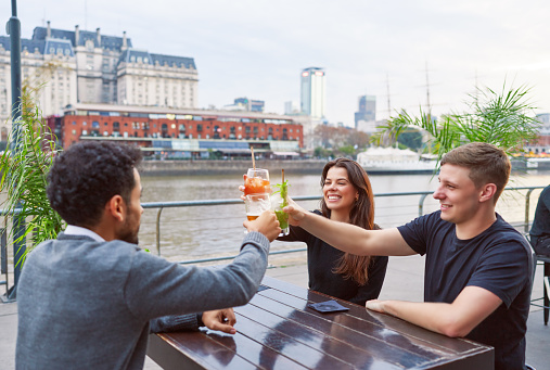 three latin american young adults friends sitting in a pub restaurant toasting happily outdoors
