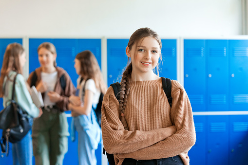Teenage girl standing with arms crossed in front of blue lockers at school and smiling at camera while her friends talking in the background.