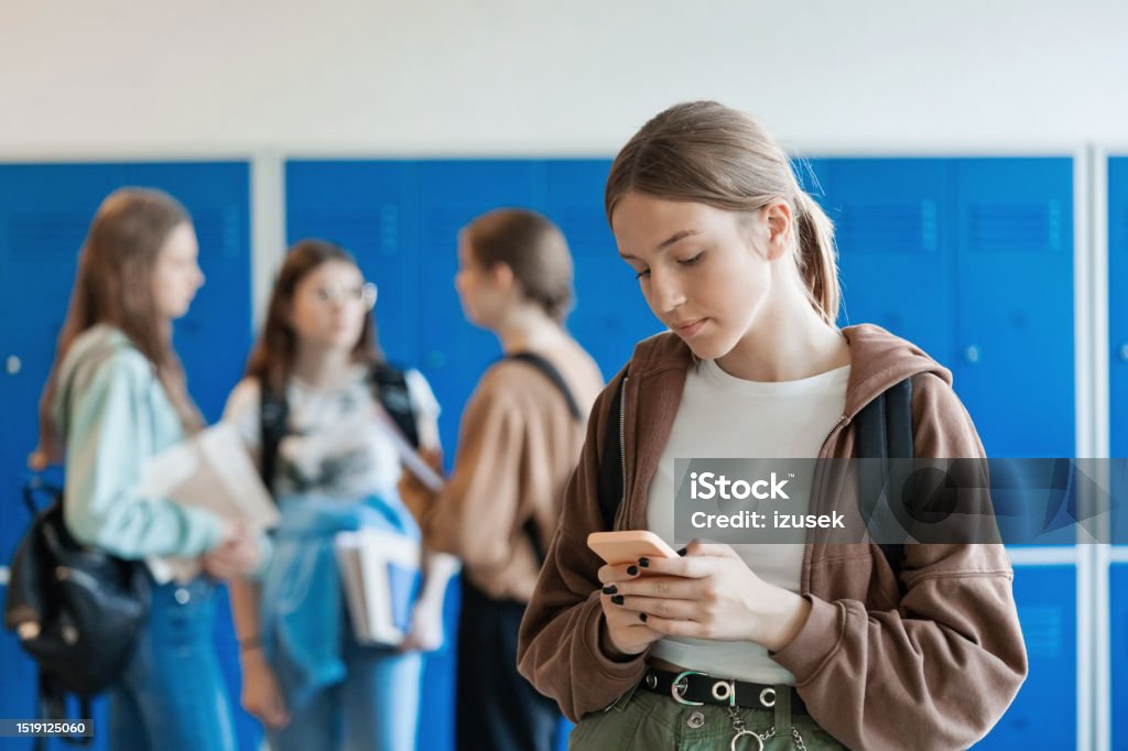 Portrait of female high school student using phone Teenage girl standing in front of blue lockers at school and using smart phone while her friends talking in the background. 14-15 Years Stock Photo