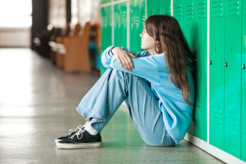 Side view of sad female high school student sitting on floor next to lockers at school and looking away.