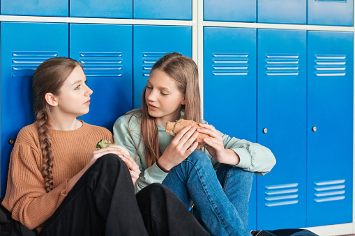 Two female high school students sitting on floor in school corridor against blue lockers and eating sandwiches.