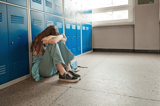 Side view of sad female high school student sitting on floor next to lockers at school.