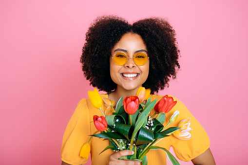 Happy lovely brazilian or african american woman with curly hair, with sunglasses, in yellow dress, holds bouquet of colorful tulips, received gift, looks at camera, smile, isolated pink background
