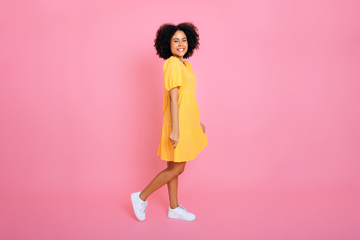 Full length photo of a happy joyful gorgeous trendy brazilian or african american curly woman, dressed in yellow sundress, looking at camera, posing, smiles friendly, stand on isolated pink background
