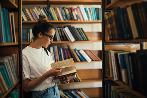 Teenage girl standing in library between bookshelves and reading book. She looks very pensive and cute