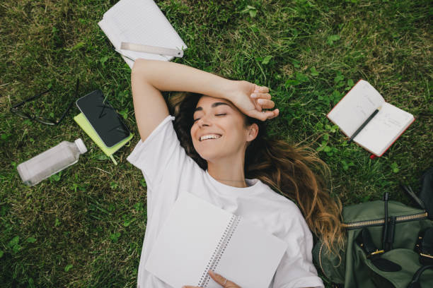 Young woman student studying using her books, laptop and notebook, sitting on grass.