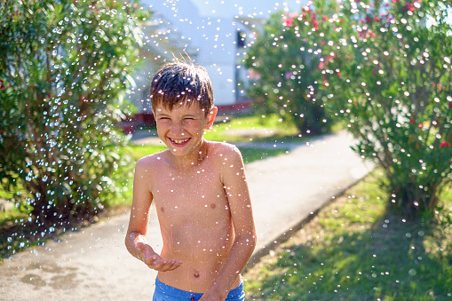 Cute boy in trunks spending fun time outdoors, splashing water in backyard on a hot summer day