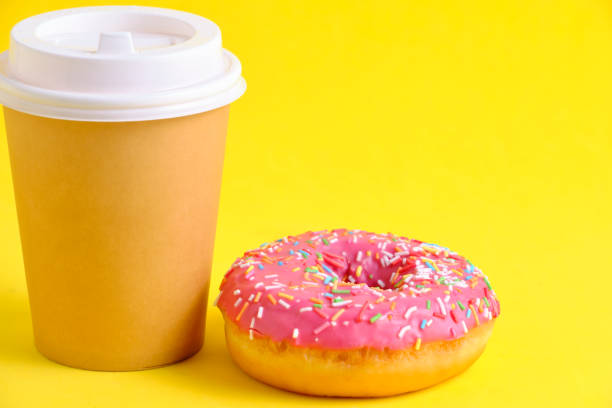 istock Disposable coffee cup and doughnut with pink icing yellow background. 1519183202