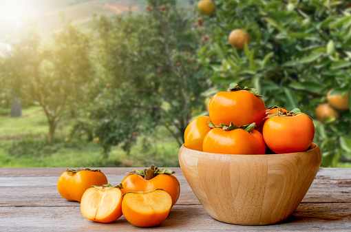 Persimmons or Persimon fruits in wooden bowl on old wooden table with persimmon tree plantation background.
