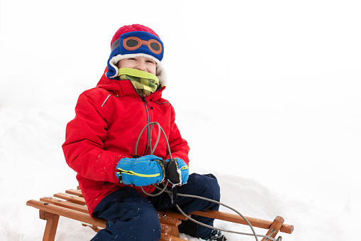 Little boy sitting on an old wooden sledge on a cold winter day with snow.